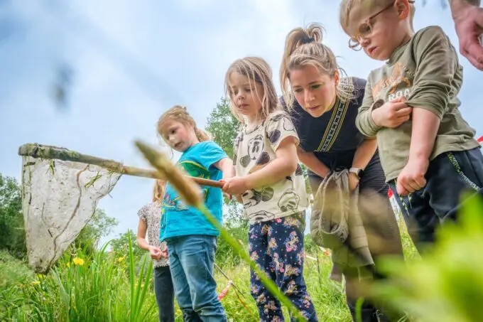 Kinderen en ouder staan langs slootkant met schepnet in water te kijken