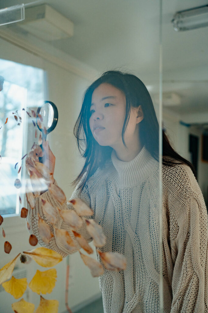 Portret van workshop docent Ying Yi Wu met vergrootglas voor een raam.